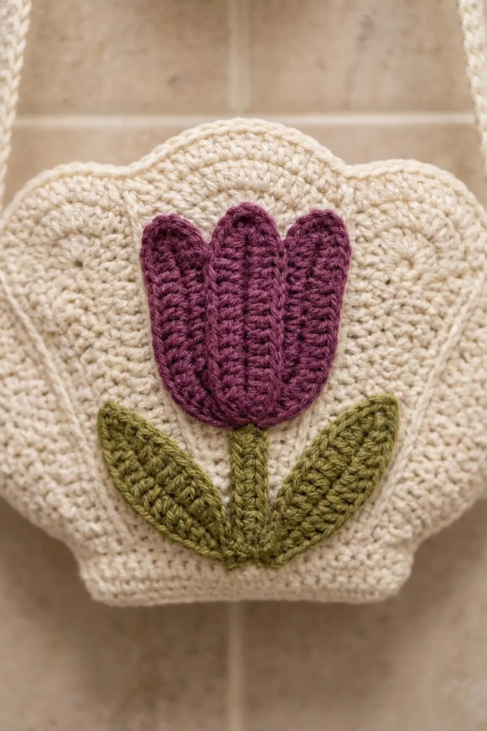 Cream crocheted cushion with burgundy tulip and green leaves appliqué against a tiled wall.