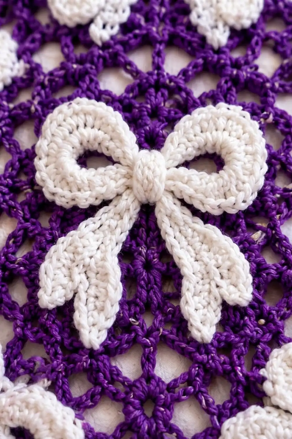 A white crocheted bow decorates the center of a purple and white lacy crocheted fabric square.