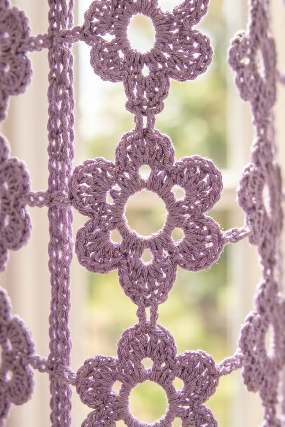 Purple crocheted lace with flower-shaped openwork motifs arranged in vertical rows, backlit with blurred foliage.