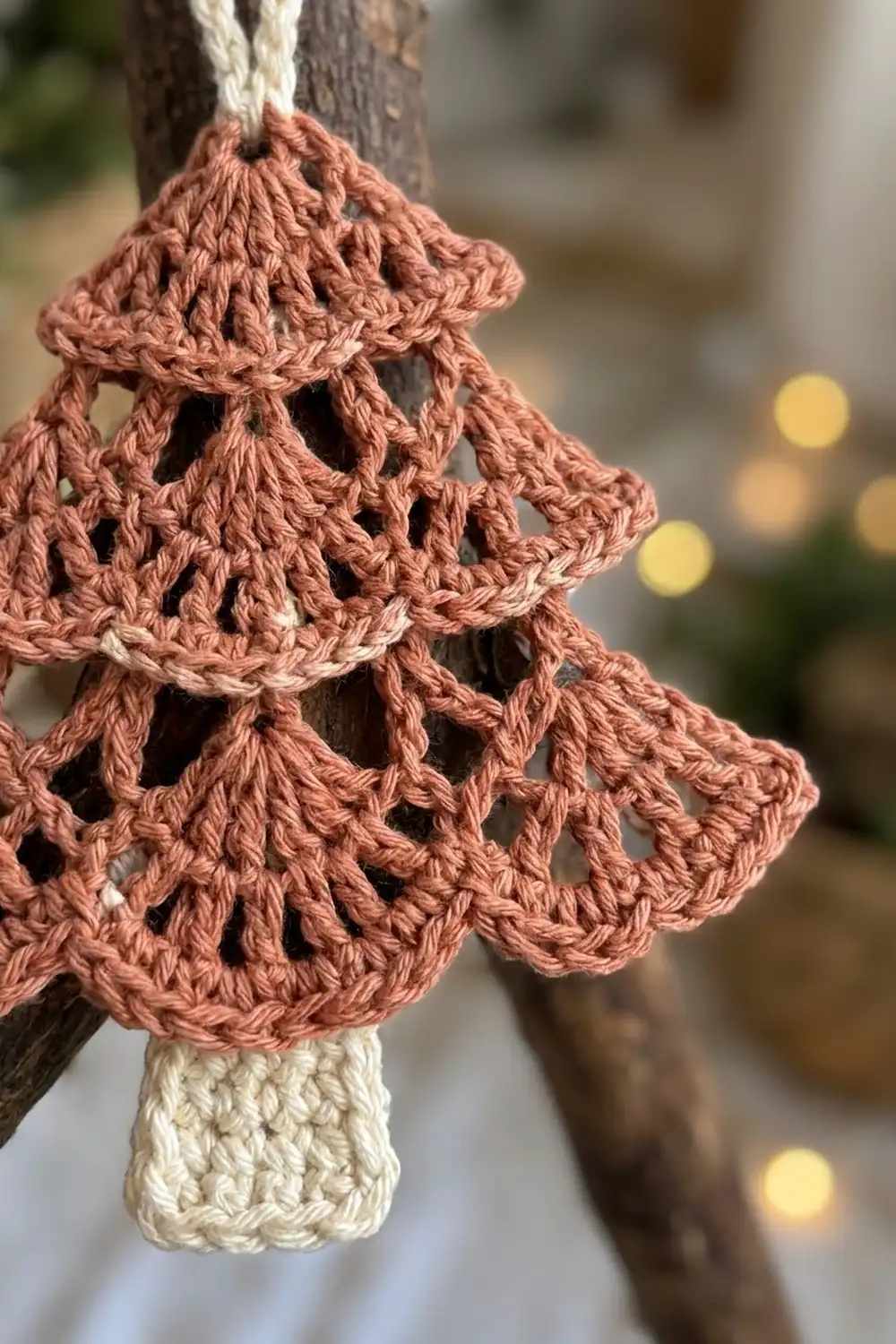 Dusty rose crocheted tree ornament with cream trunk hanging from a cord against soft golden bokeh background.