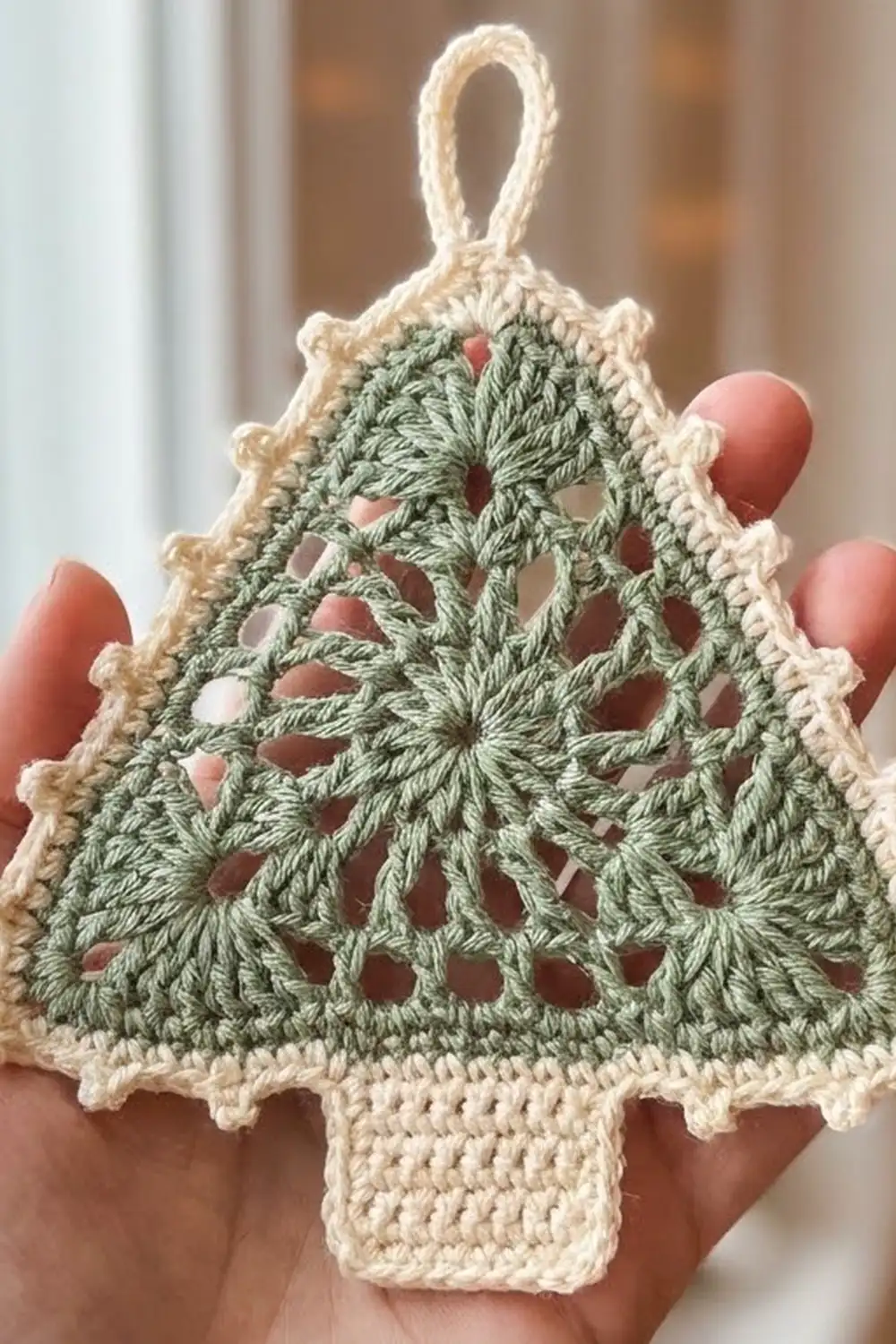 A sage green crocheted Christmas tree ornament with cream lace edging held in a hand.