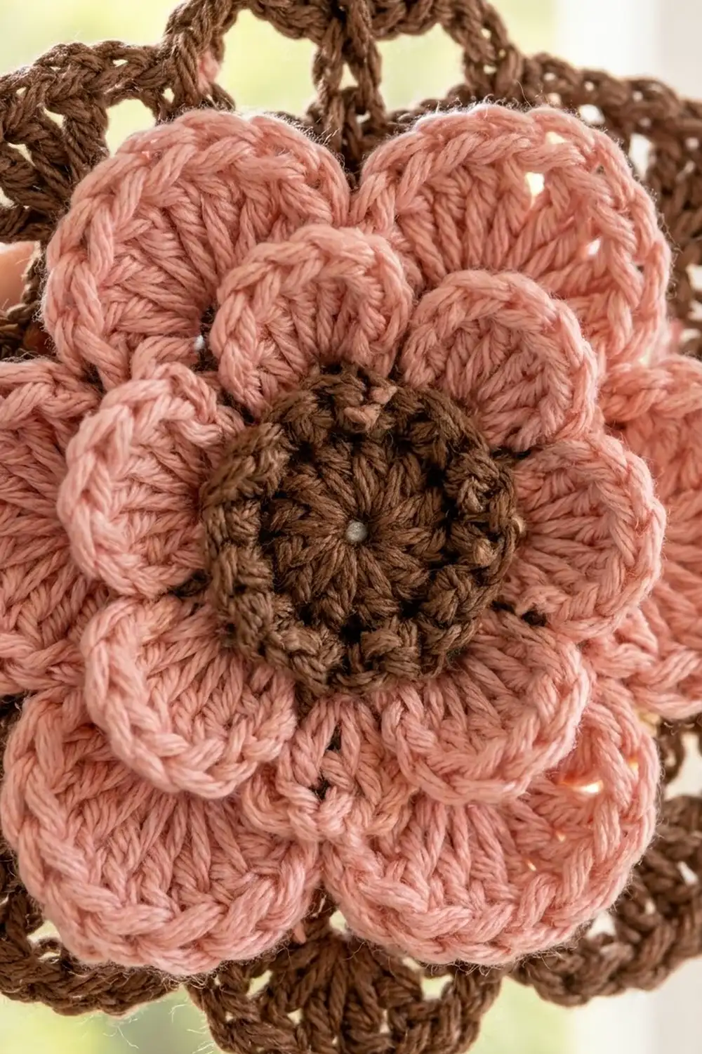 Close-up of a pink crocheted flower with brown center and stem against a soft yellow background, showing detailed yarn texture.
