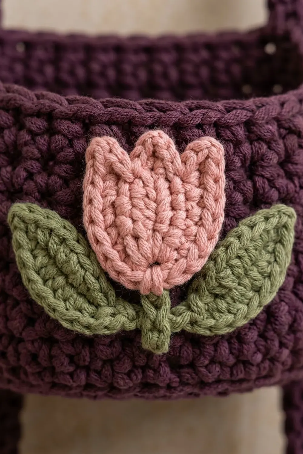 Close-up of a crocheted pink tulip with green leaves on a dark purple textured crochet fabric.