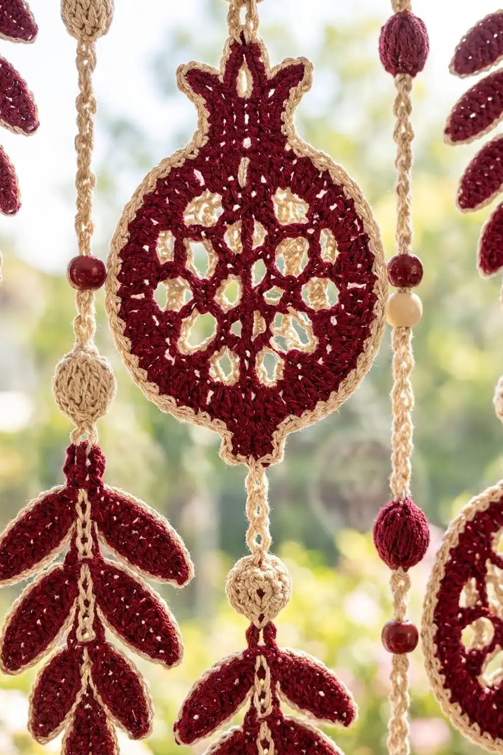 Handcrocheted deep burgundy pomegranate with tan lace details hangs from beaded chains against a blurred green backdrop.
