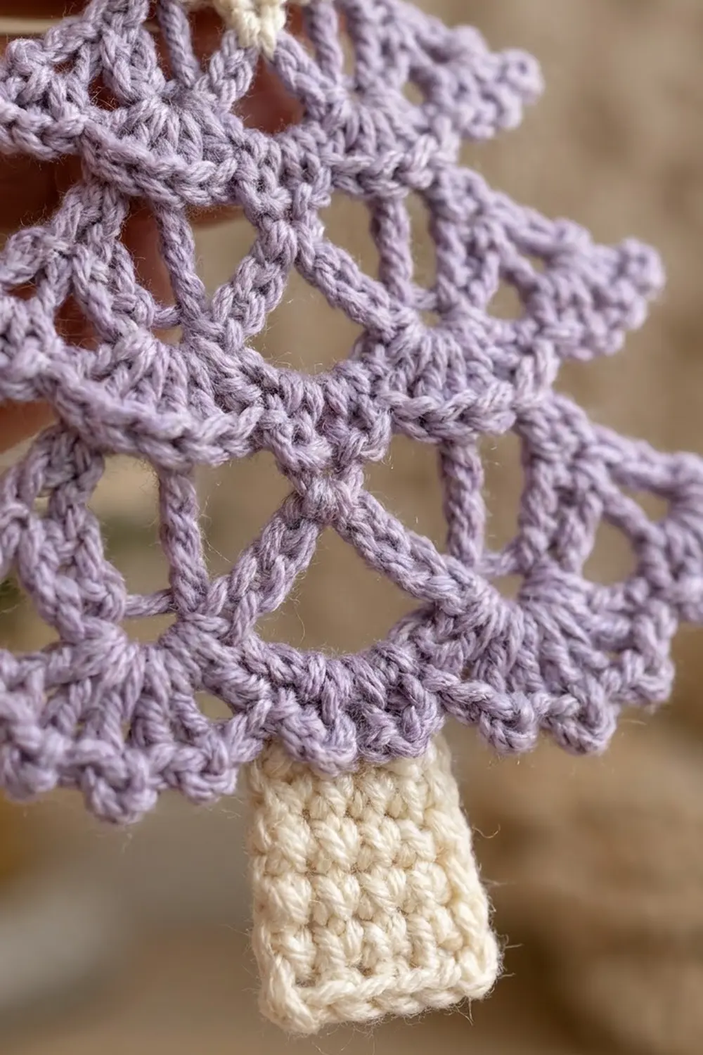 Delicate lavender crocheted lace with open stitches and scalloped edges attached to a cream-colored base against a soft background.