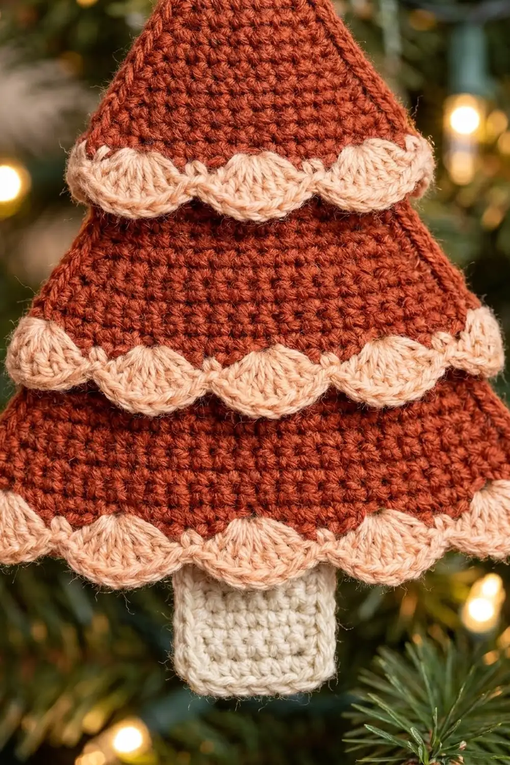Crocheted Christmas tree ornament in rust red with cream-colored scalloped trim and trunk, against festive holiday lights.