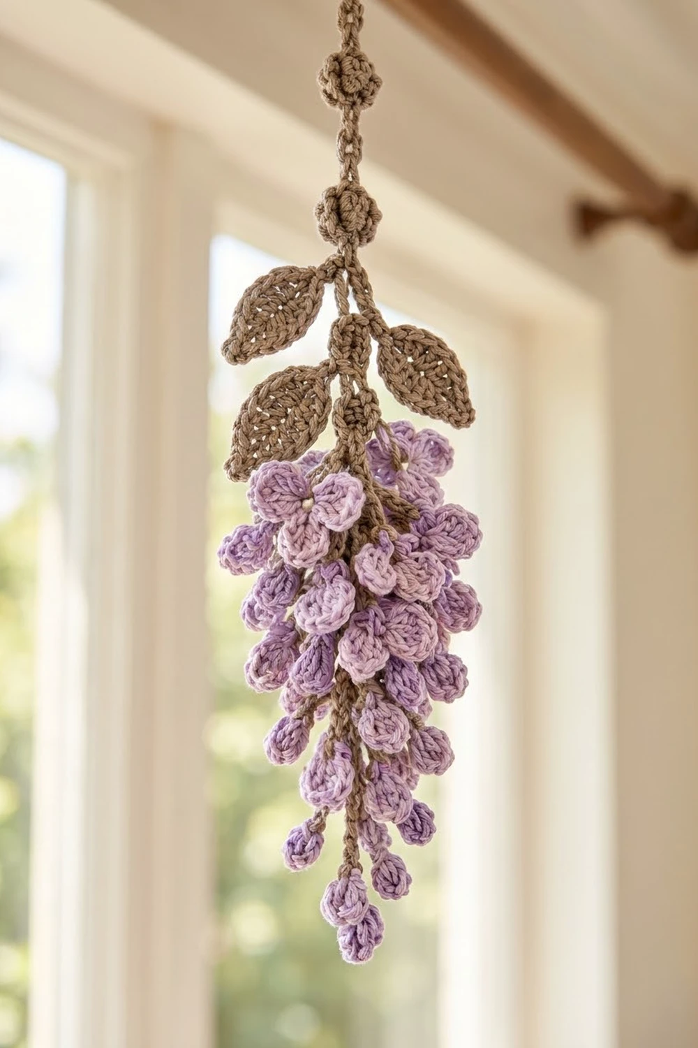 Crocheted wisteria hanging ornament with purple flowers, tan leaves, and braided cord suspended in a bright window.