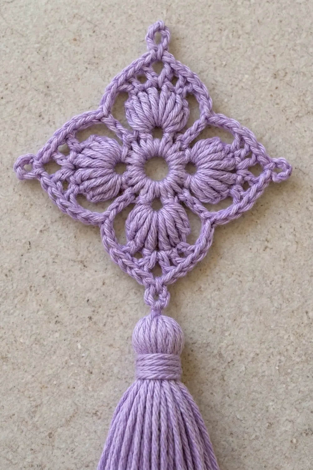 A lavender crocheted ornament with a four-pointed star design and tassel hanging against a beige textured background.
