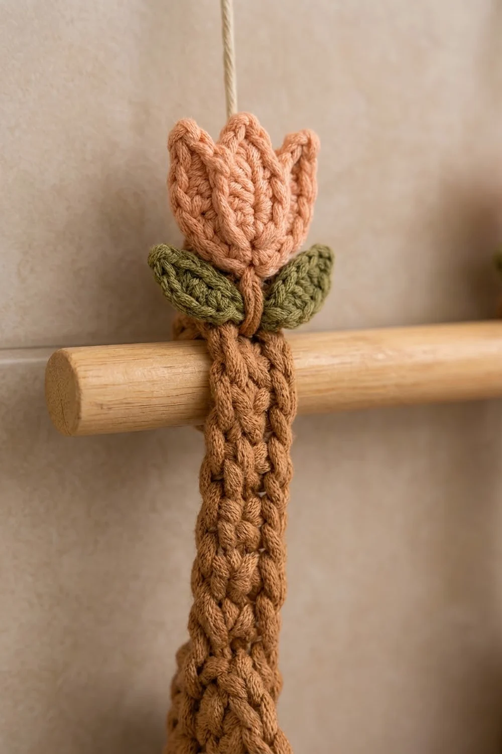 A crocheted peach tulip with olive leaves and brown stem wrapped around a wooden dowel hangs from cream rope.