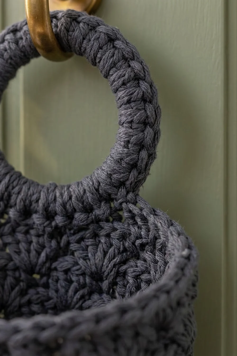 Charcoal-black crocheted rope with detailed stitch texture looped through a brass ring against a soft green background.