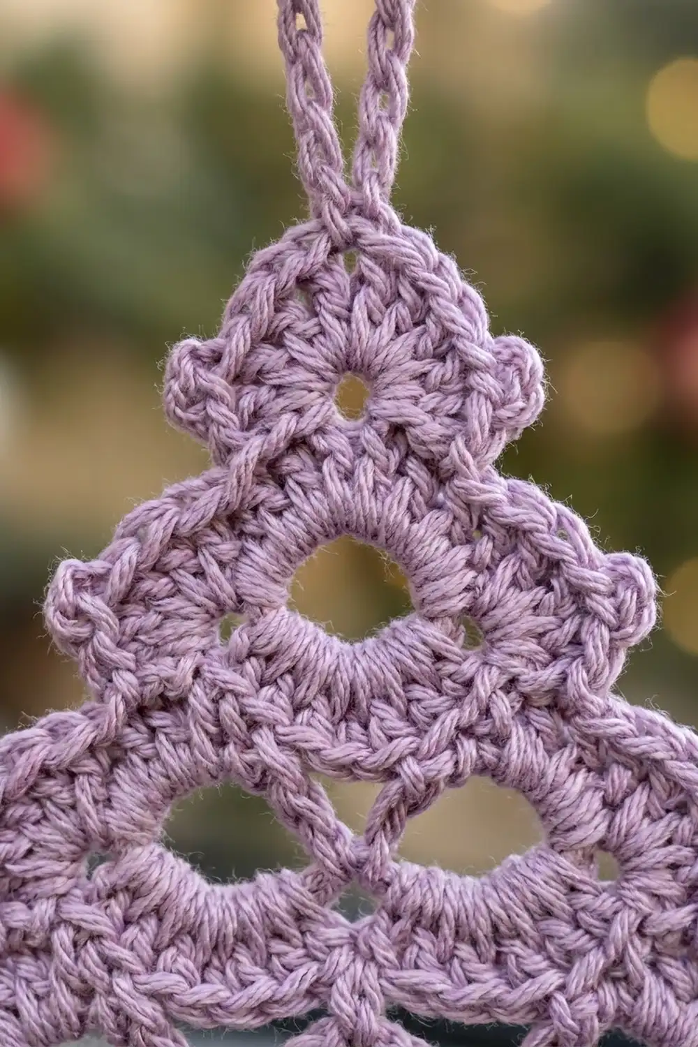 Lavender crocheted geometric pendant with openwork pattern and braided cord hanger in soft outdoor setting.