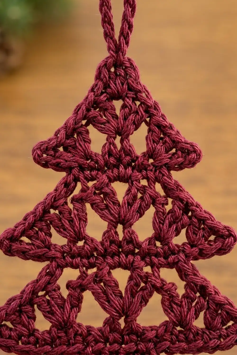 Burgundy crocheted triangle with intricate openwork geometric pattern and braided loop, photographed against a soft blurred tan background.