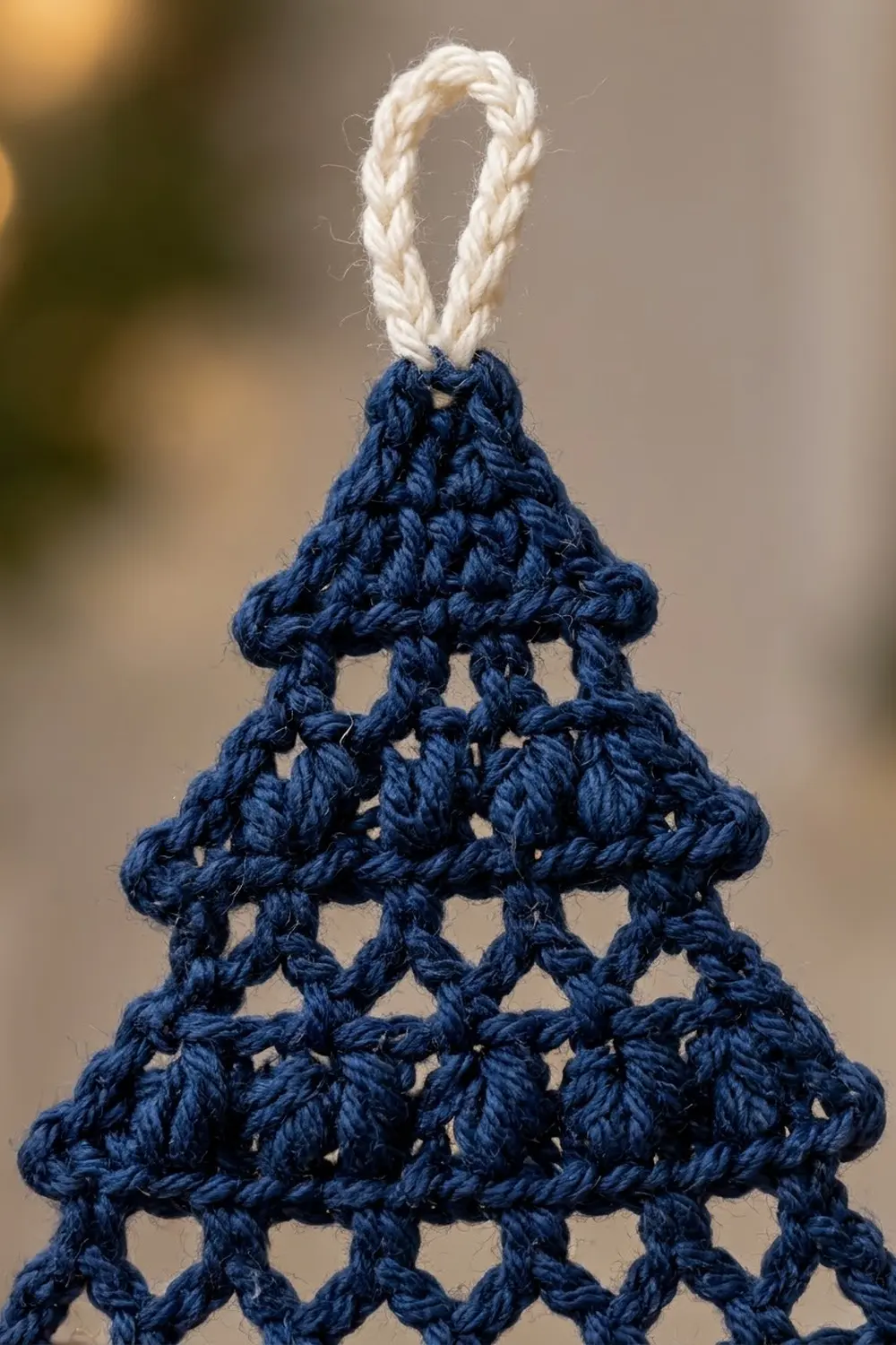 Crocheted navy blue Christmas tree ornament with lattice stitch pattern and cream rope loop hanging against a blurred warm background.