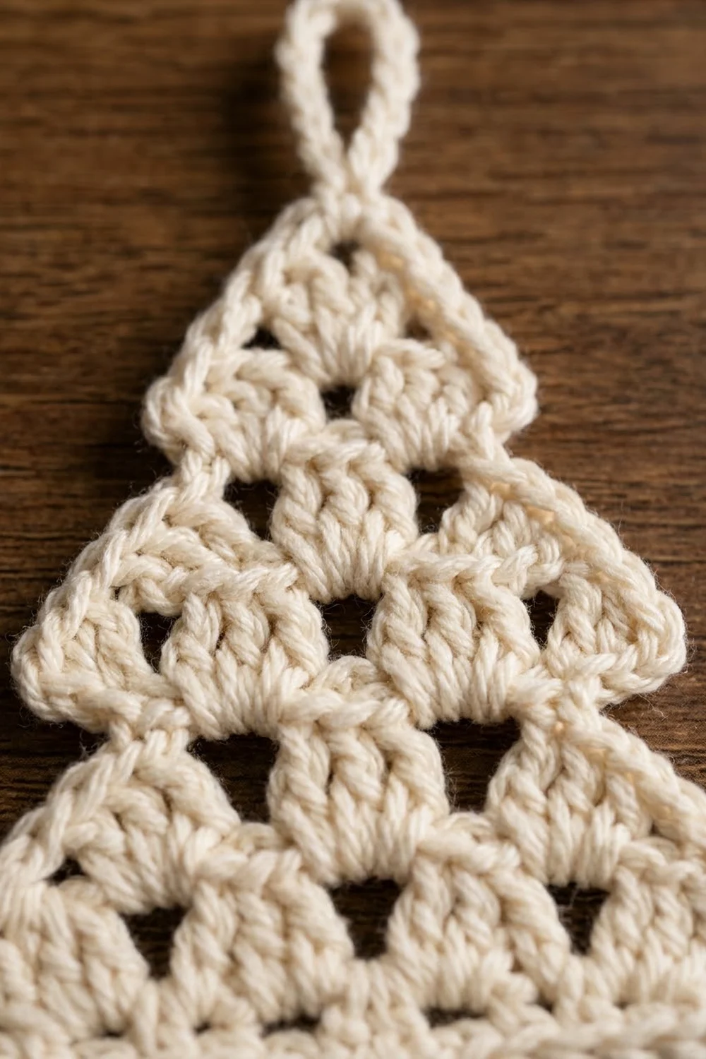 Cream crocheted Christmas tree ornament with bumpy textured stitches and hanging loop on dark wood.