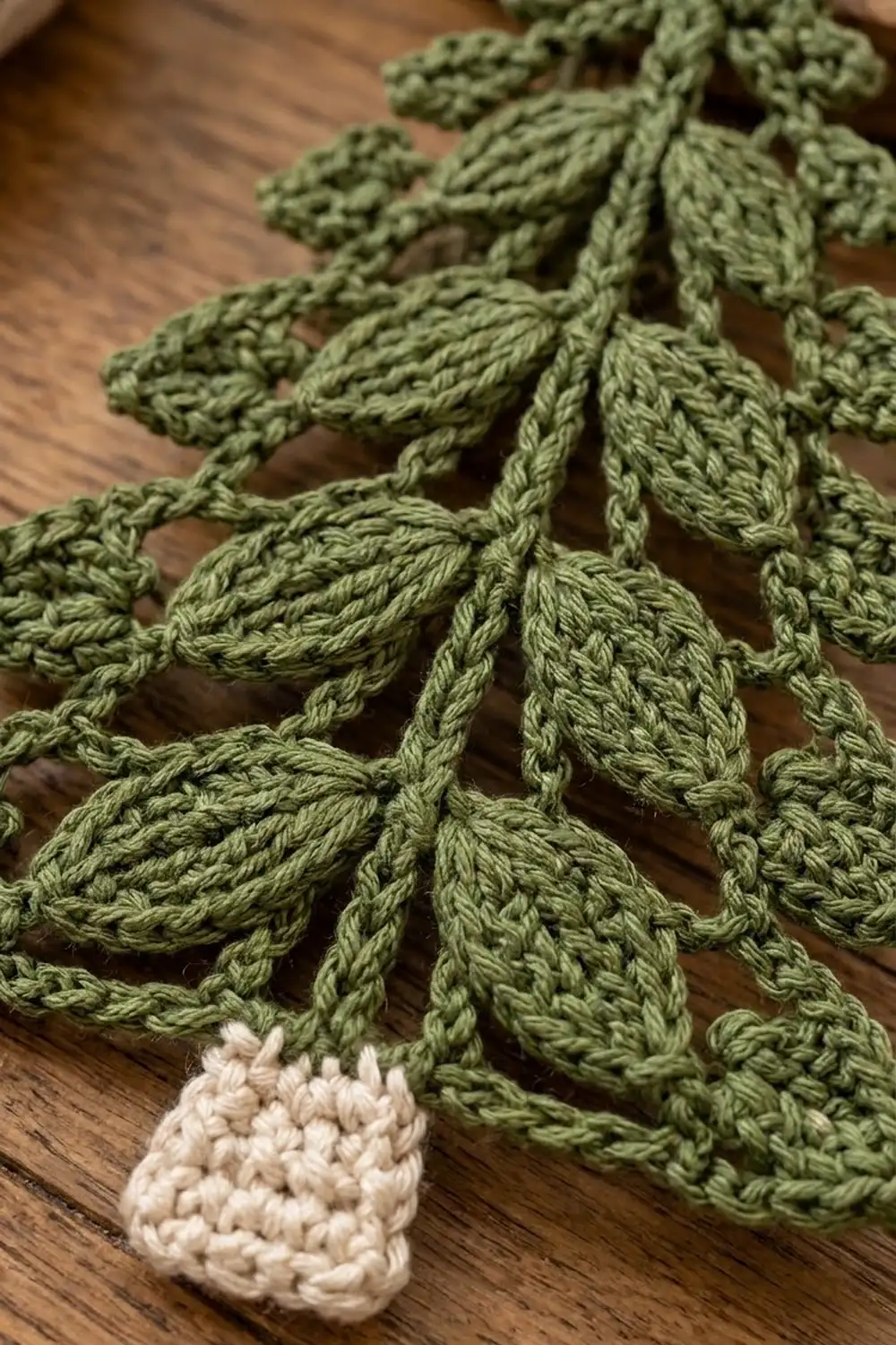 Crocheted fern-like leaves in sage green with a cream bobble detail on a warm wooden surface.