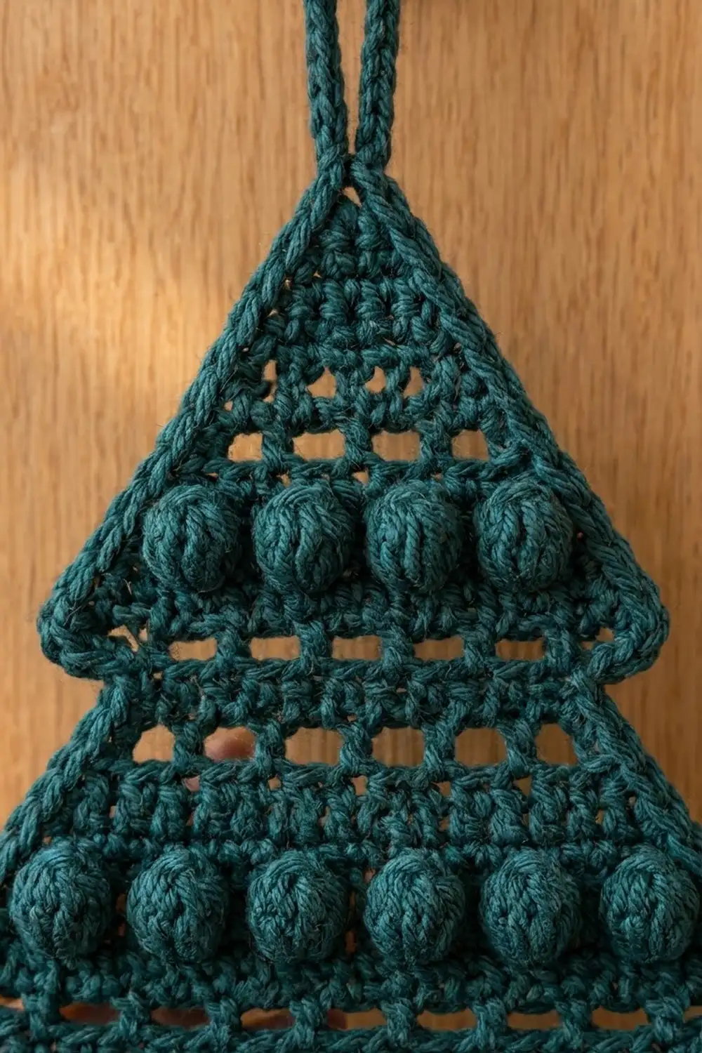 Teal crocheted triangle ornament with openwork lattice pattern and popcorn bobbles against wooden backdrop.