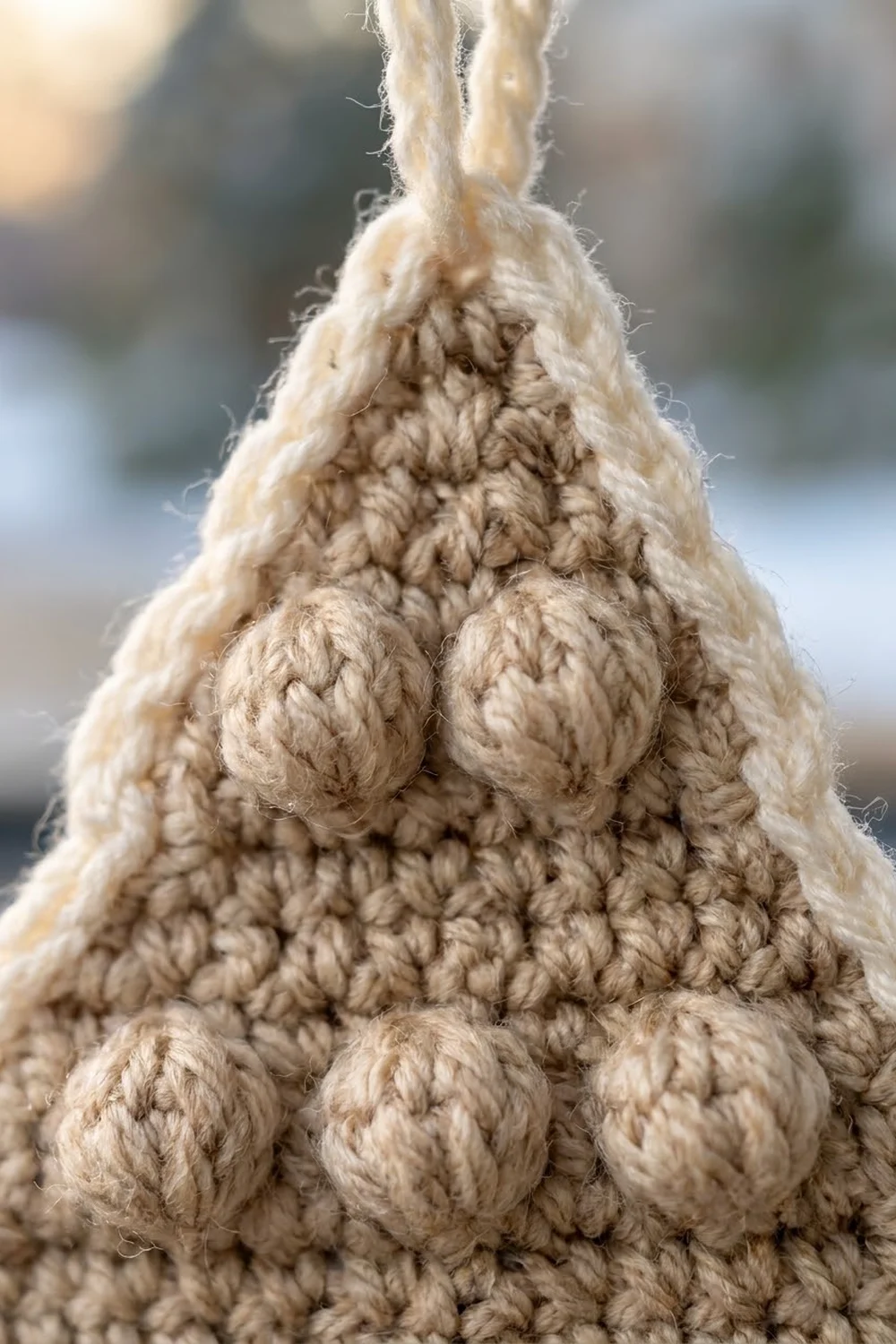 Close-up of a cream crocheted pear ornament with bobble stitch texture, tied with a cord against a blurred background.