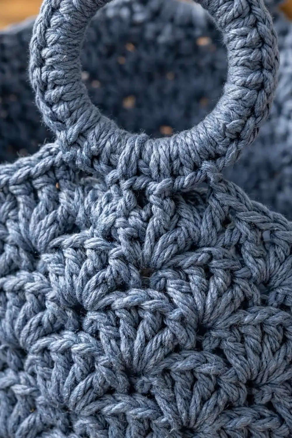 Gray crocheted fabric showing detailed stitch patterns with a circular opening, captured in close-up against a warm, blurred background.