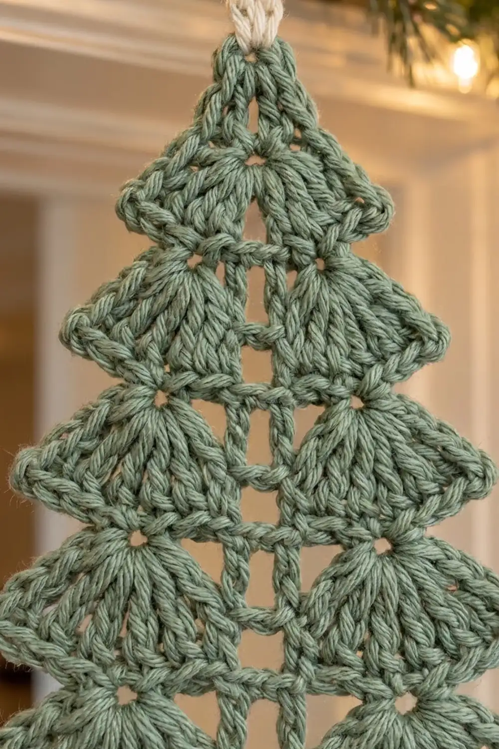 Sage green crocheted Christmas tree with cream tie displays intricate stitchwork in tiered rows against a warm, golden background.