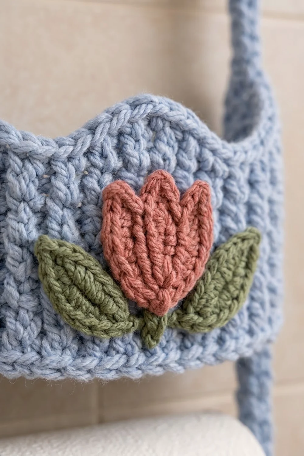 A crocheted red flower with green leaves sits on a light blue knitted surface against a soft beige background.