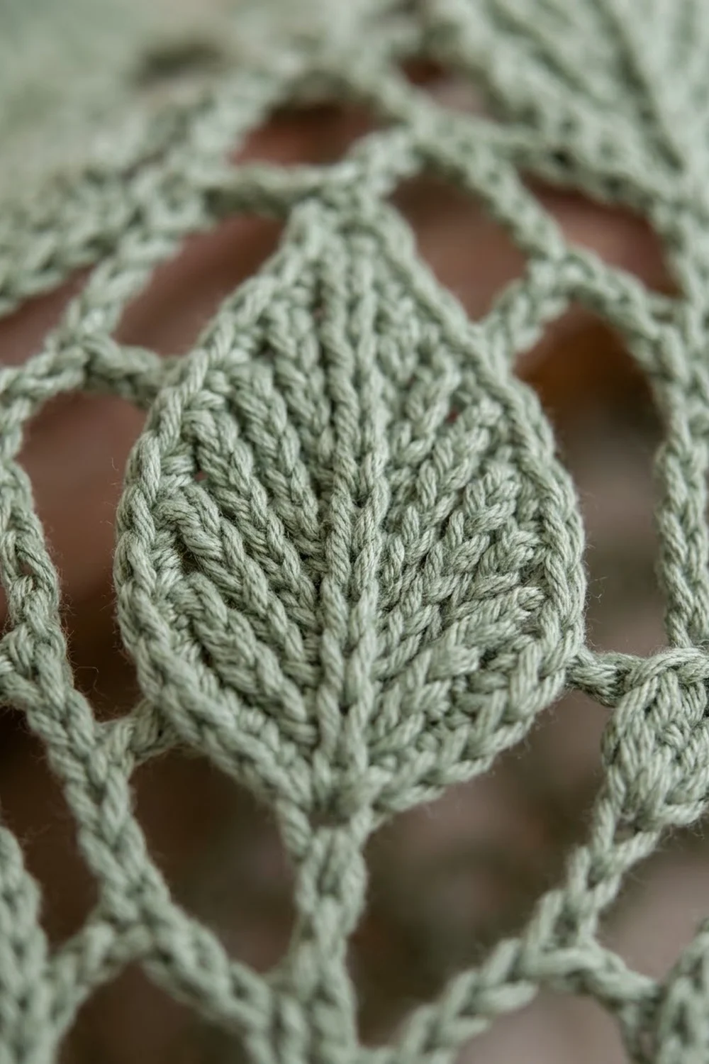 Sage green crocheted lace featuring an intricate cable mesh pattern with an open, delicate texture over a blurred neutral background.
