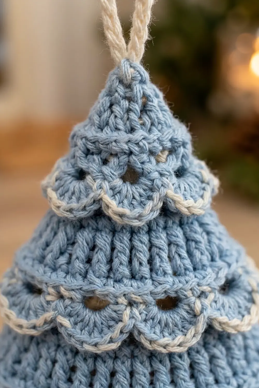 Handmade crocheted Christmas tree ornament in dusty blue with cream trim, displaying intricate stitch detail on blurred outdoor background.