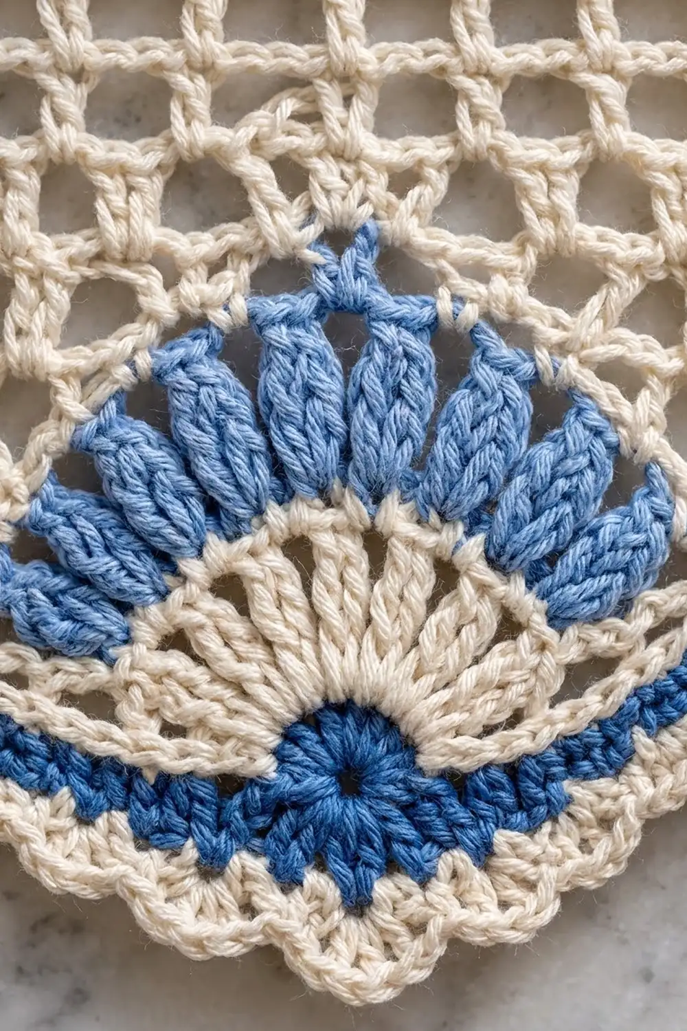 Close-up of a crocheted motif featuring blue and cream yarn in a flower-like pattern on a net background.