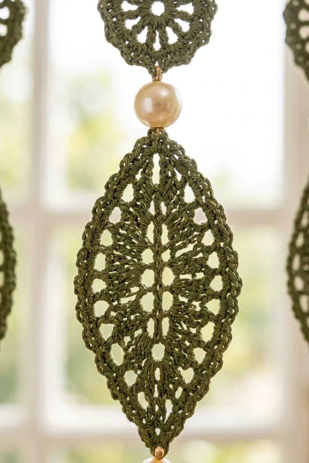 Olive-green crocheted ornament with cream pearl focal bead and delicate openwork pattern, hung at a bright window.