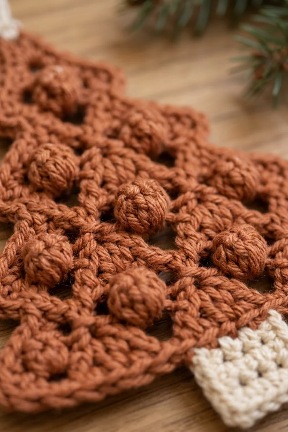 Close-up of rust-brown crocheted popcorn stitch with cream detail on wooden surface beside evergreen foliage.