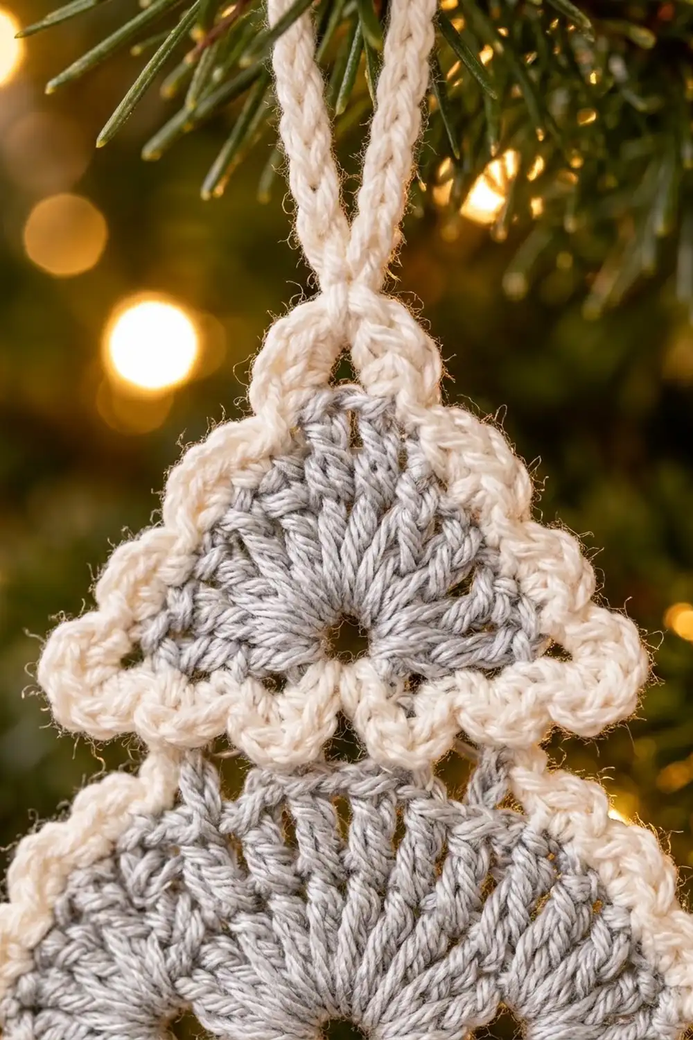 Crocheted tree ornament with stacked cream and gray layers hanging from white rope with evergreen and bokeh lights.