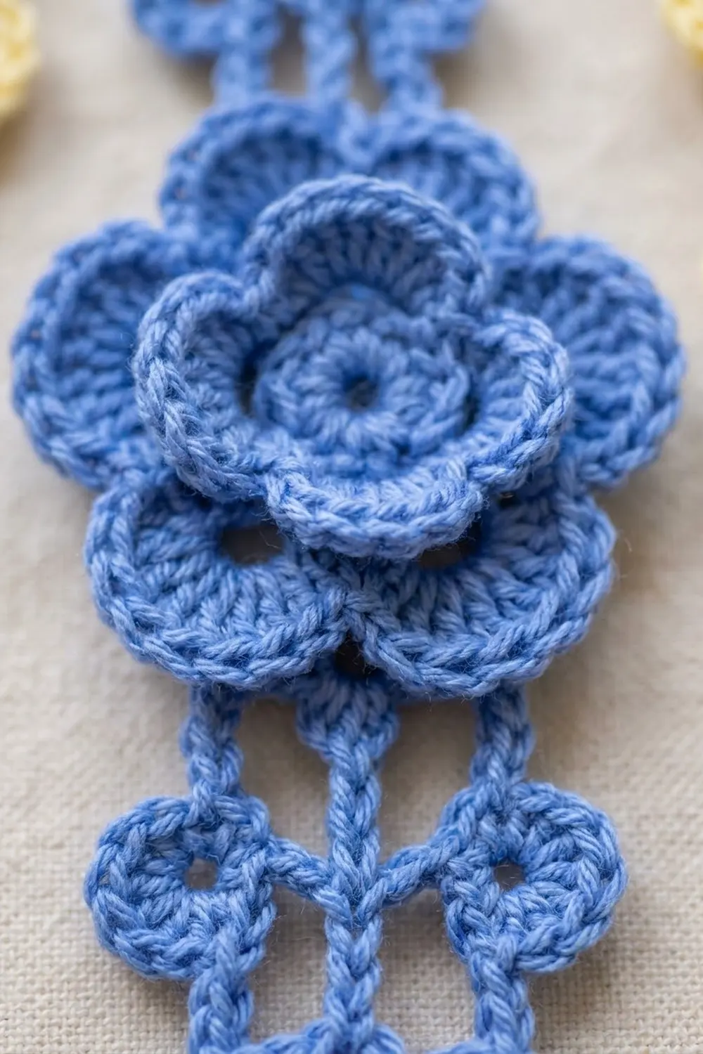 A blue crocheted flower ornament with layered petals and decorative hanging strings on a beige fabric background.