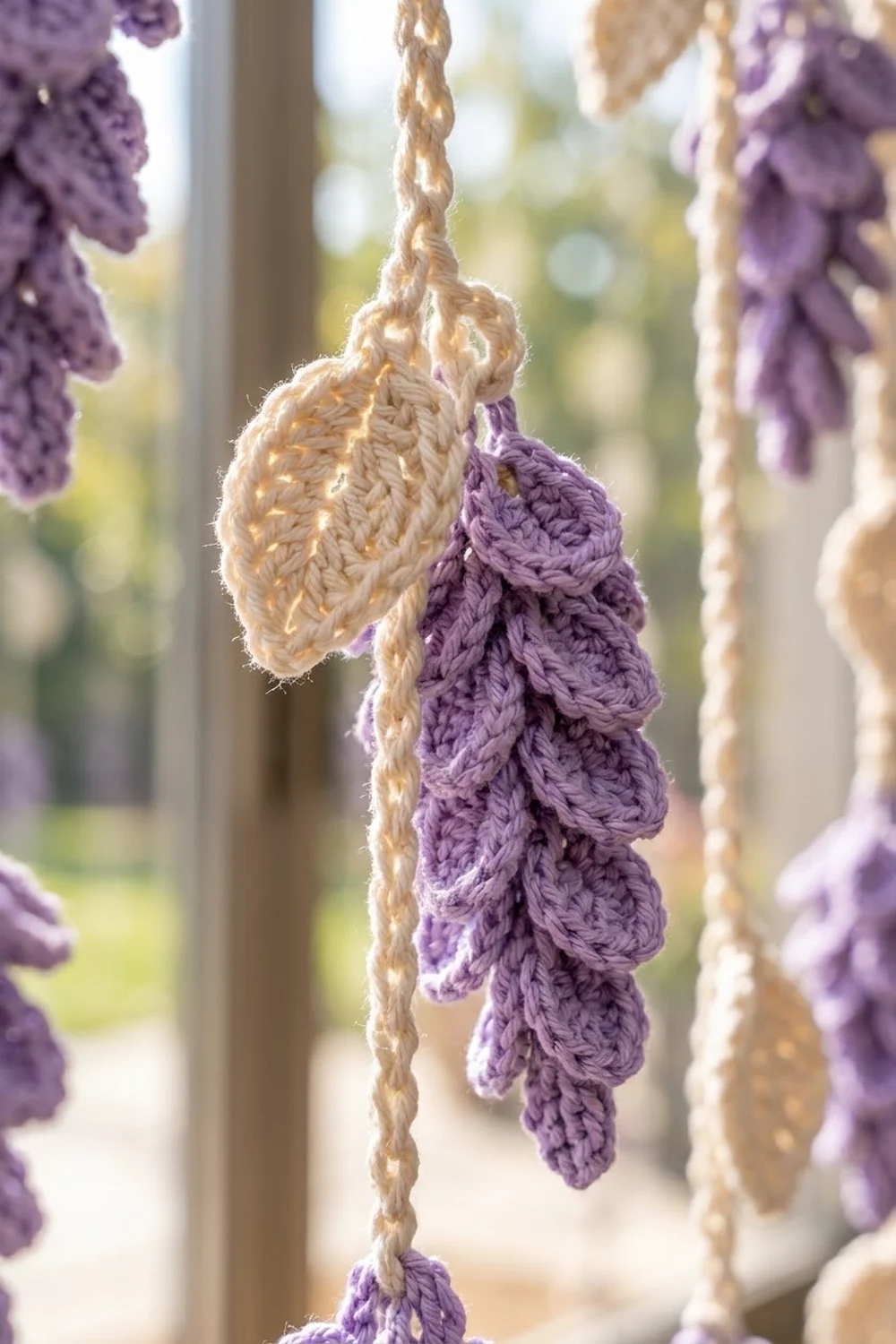Crocheted wisteria flowers in purple and cream hang from braided jute cords near a sunny window.
