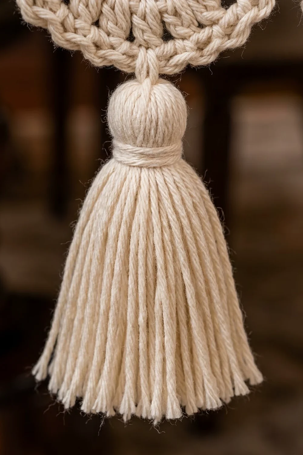 A cream-colored rope tassel with braided loop and fringe skirt hanging against a blurred brown background.