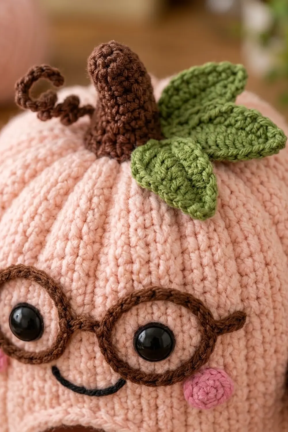 A crocheted pink cactus toy with brown details, black eyes, and green leaves against a blurred background.
