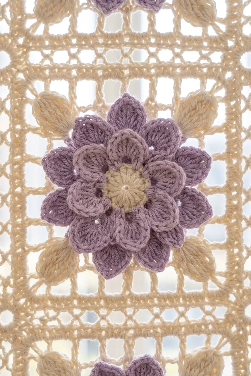 A crocheted purple flower with cream center appliquéd onto a beige openwork crochet background panel.