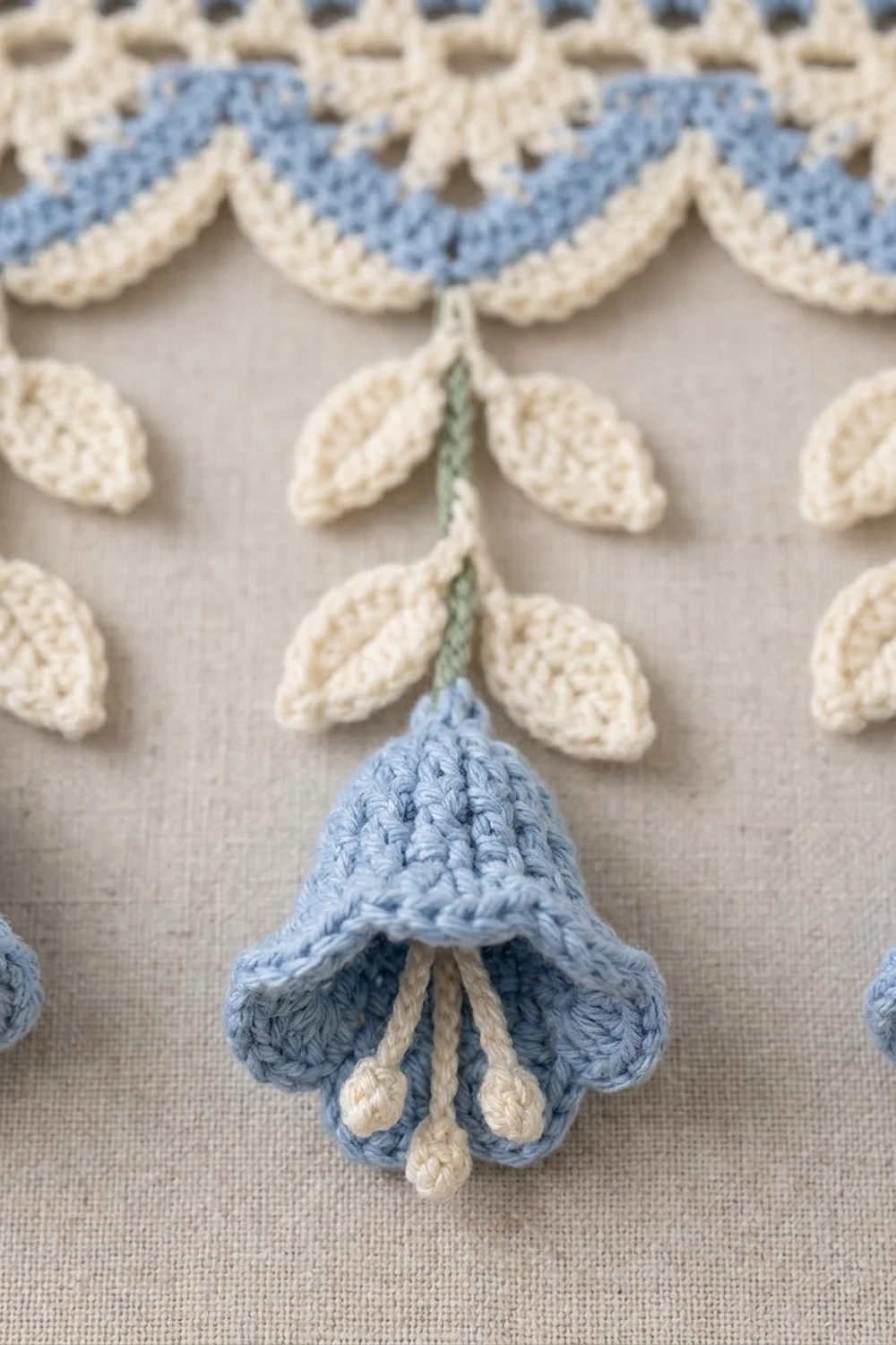 Crocheted bell-shaped flowers in blue and cream with decorative scalloped leaves arranged on beige fabric.