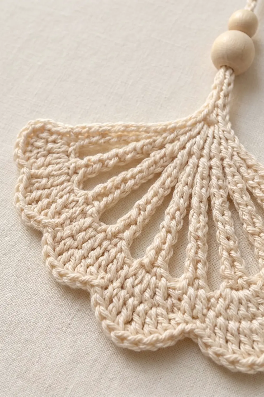 Cream-colored crocheted leaf ornament with wooden beads attached to a string, displayed on white linen fabric.