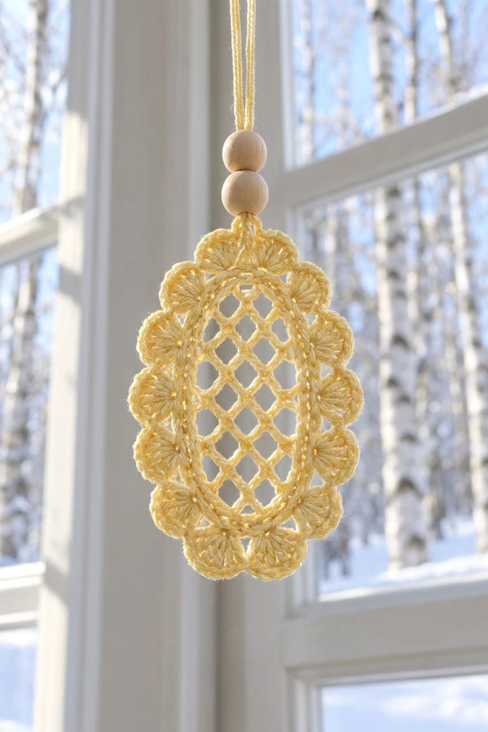 Golden crocheted ornament with diamond lattice pattern hanging in a sunlit window with snowy trees visible outside.