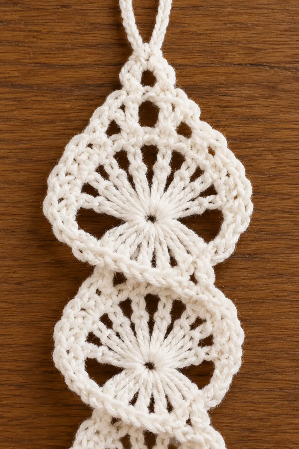A white macramé ornament with two circular medallions featuring radiating knots, hanging against a wooden surface.