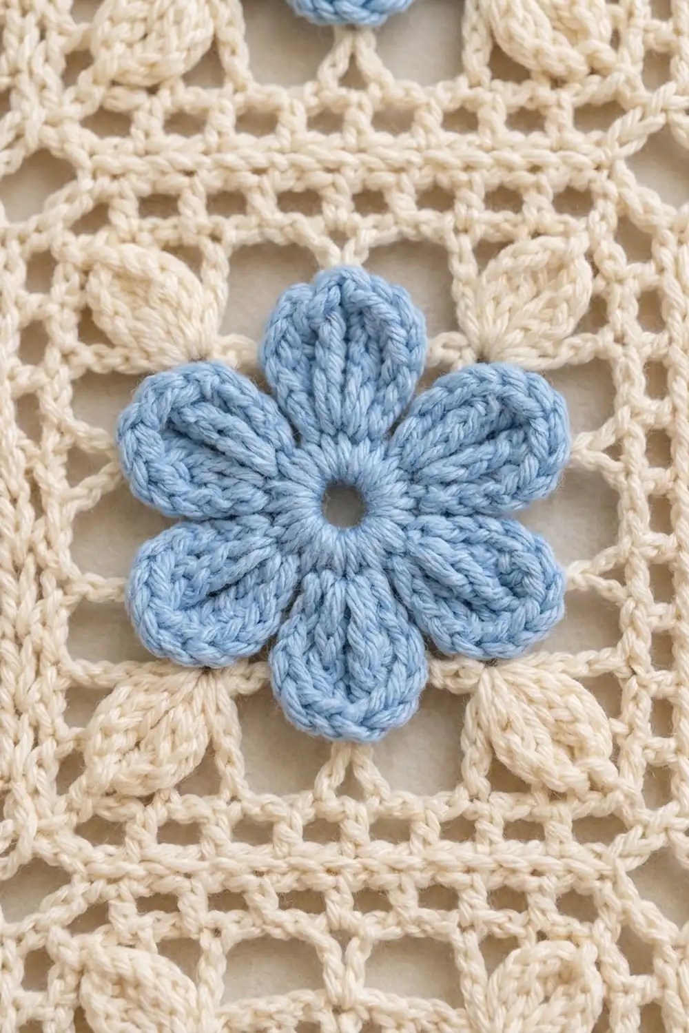 A light blue crocheted flower with six petals sits centered on a cream-colored lacy crochet background.
