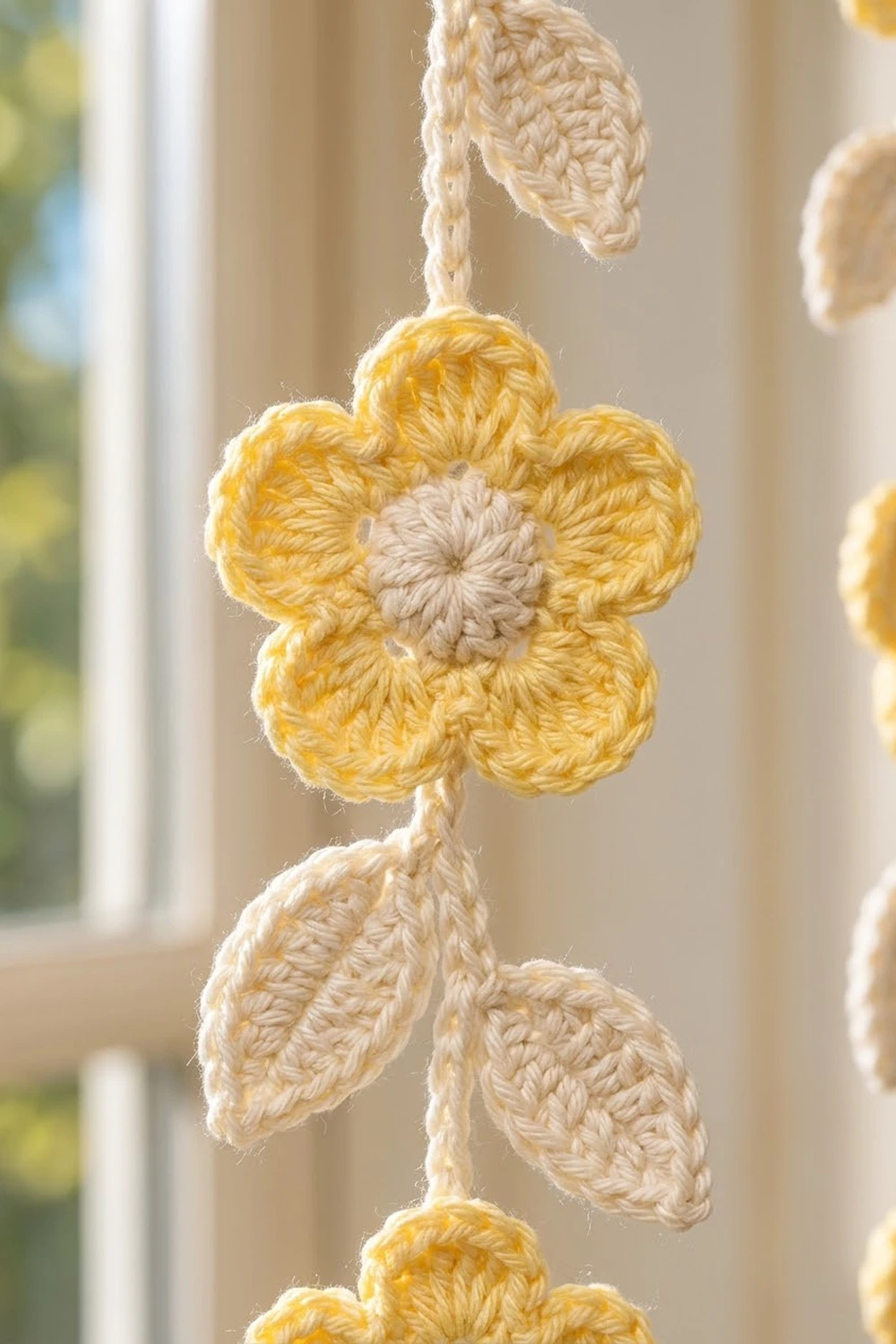 Crocheted yellow flowers with beige centers and leaves hanging by twine near a sunny window.