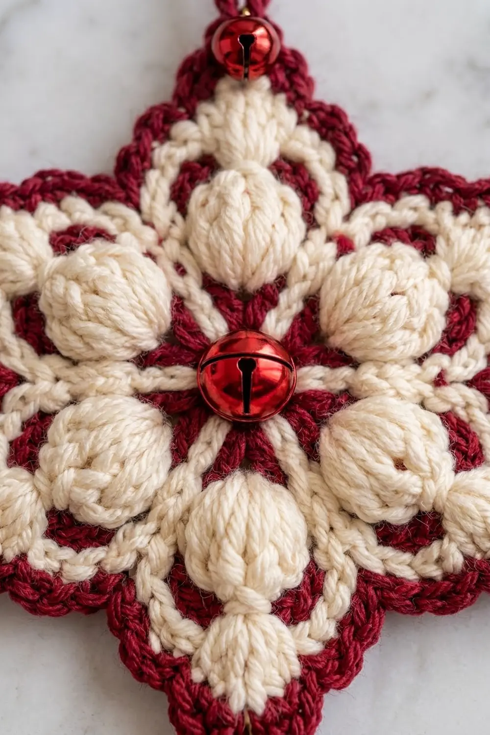 A red and cream crocheted snowflake ornament with metallic red jingle bells hanging against a white background.