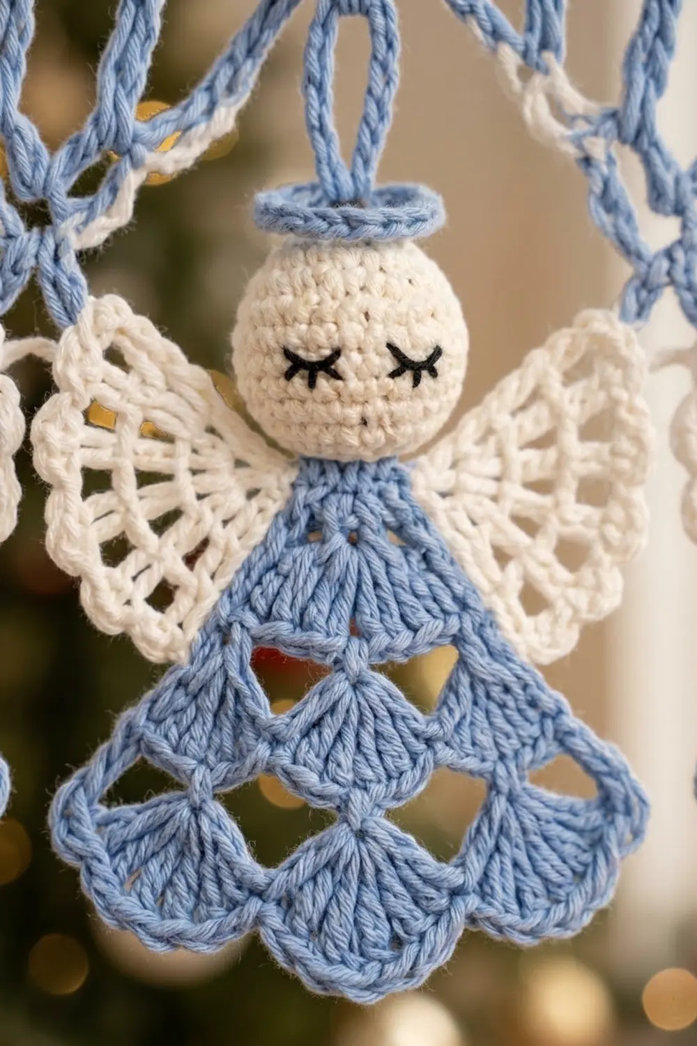 A handmade crocheted angel ornament with a cream head, blue dress, white lace wings, and closed eyes hanging from a blue cord.