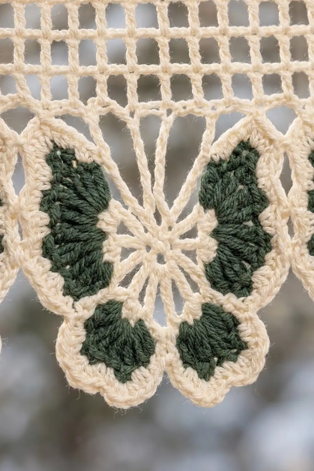 Close-up of a crocheted butterfly appliqué with cream and dark green yarn against a macramé lattice background.