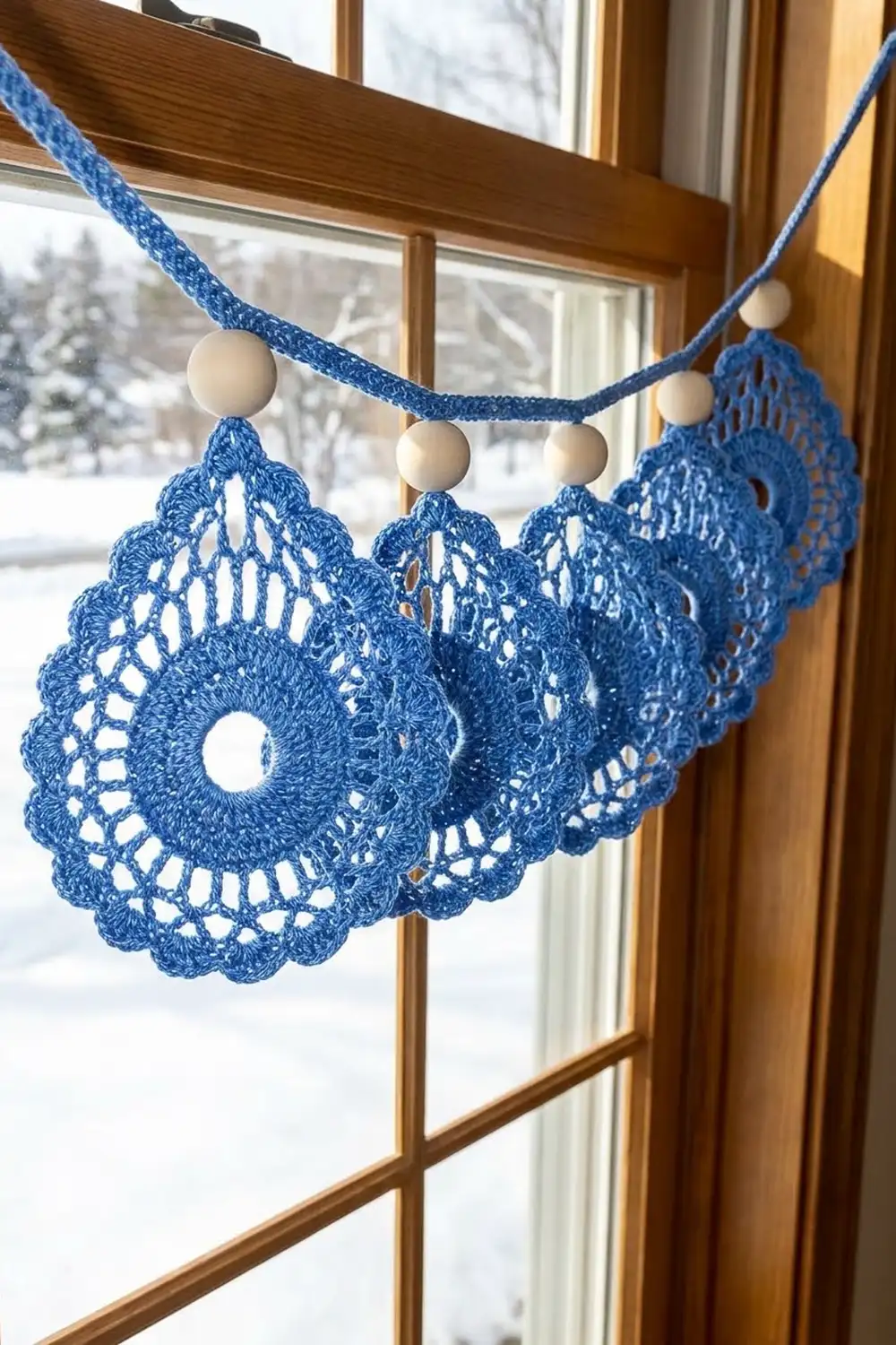 Blue crocheted doilies with wooden beads strung on blue cord hanging in a snowy window.