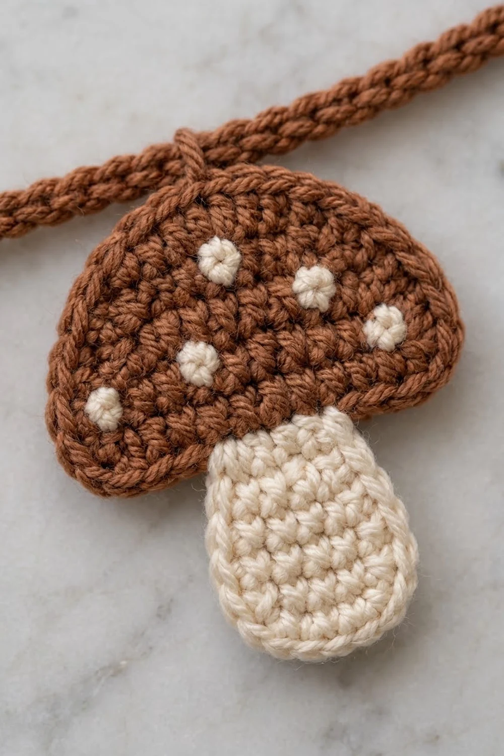 Crocheted mushroom ornament in brown and cream attached to a twisted rope cord against a marble background.