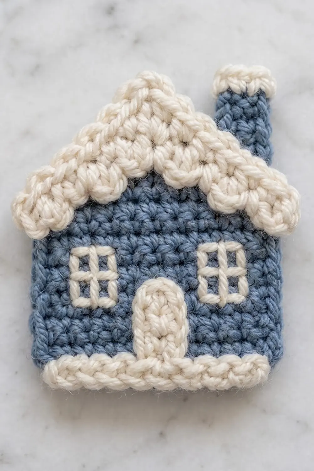 A crocheted blue house ornament with cream roof, windows, door, and chimney on marble background.
