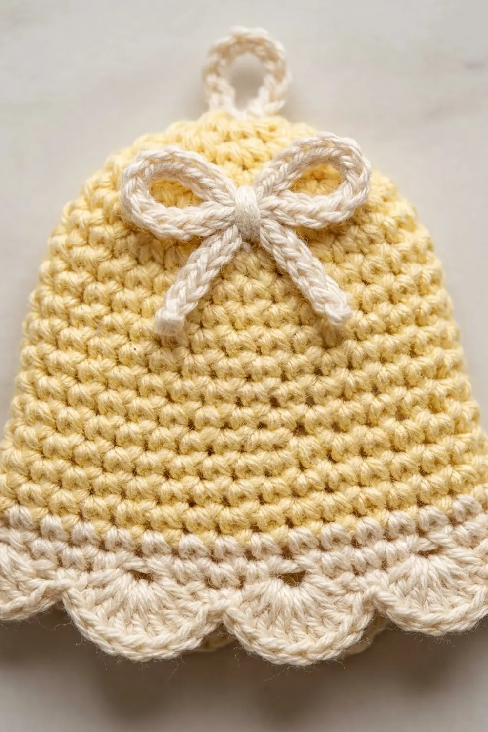 A crocheted bell ornament in cream and pale yellow with a white bow, photographed against a neutral background.
