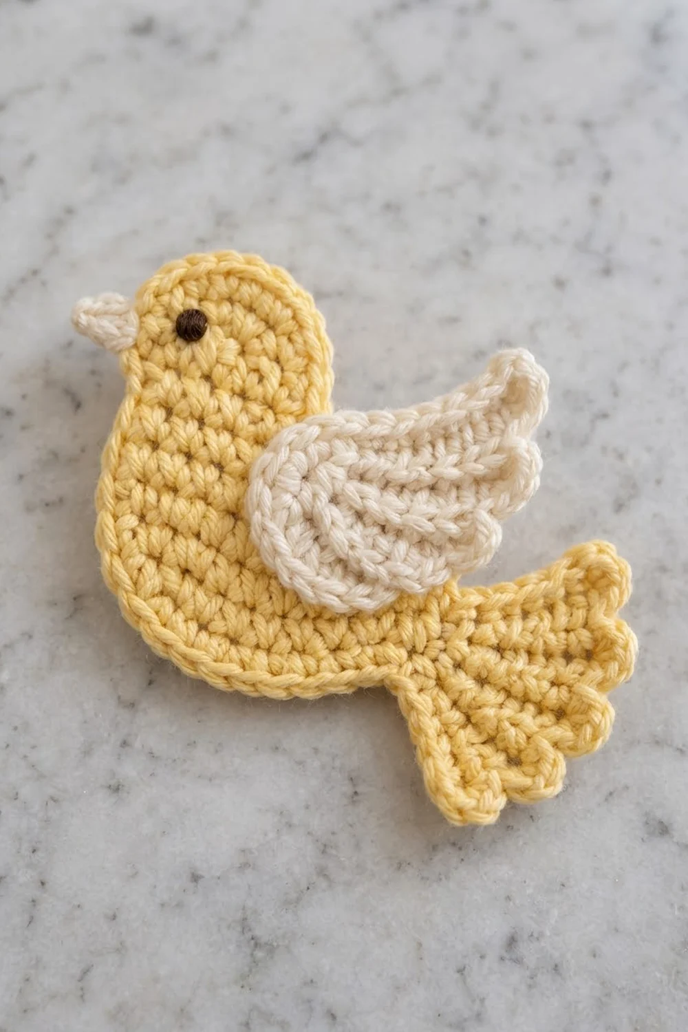 A crocheted yellow duck with cream-colored wings and a black bead eye sits on a marble surface.