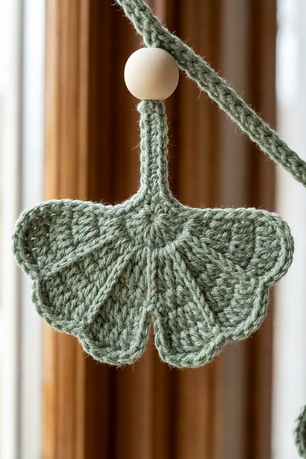 A sage green crocheted butterfly ornament with a cream bead hanging from green yarn against blurred curtains.